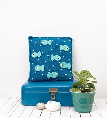 Gehaakte, donkerblauwe kussen met lichtblauwe visjes op een wit tafeltje, tegen een witte muur. Op de foto staan verder een plant in een groene plantenpot en een paar schelpen.
