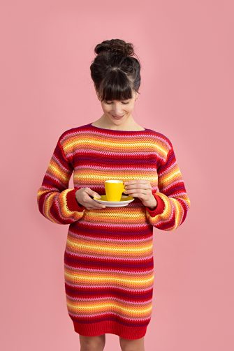 Model draagt de gestreepte jurk in de kleuren rood, oranje en geel die steeds in elkaar overlopen en houdt een geel theekopje met schoteltje vast.