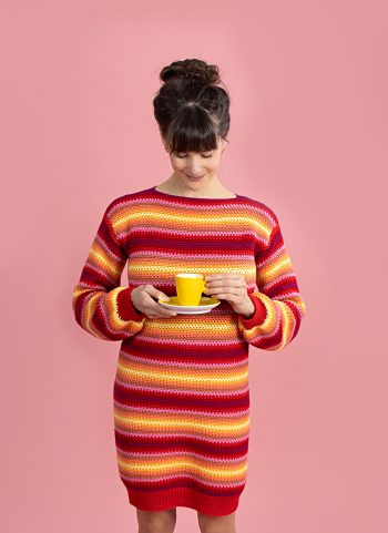 Model draagt de gestreepte jurk in de kleuren rood, oranje en geel die steeds in elkaar overlopen en houdt een geel theekopje met schoteltje vast.