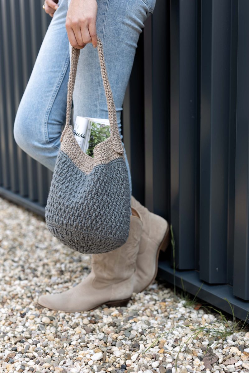 Een model houdt de gehaakte tas losjes in haar hand en leunt tegen de muur aan. Ze draagt een spijkerbroek en beige cowgirllaarzen. De tas is blauw met een grijze handel.